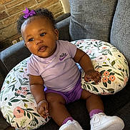 Jariya is registered to the contest to win money with this photo: baby, bow, brick_wall, casual, child, comfortable, couch, curly_hair, cute, floral_pattern, footwear, headwear, indoor, nursing_pillow, person, pillow, purple_clothing, sitting, smiling, white_sneakers