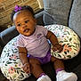 baby, bow, brick_wall, casual, child, comfortable, couch, curly_hair, cute, floral_pattern, footwear, headwear, indoor, nursing_pillow, person, pillow, purple_clothing, sitting, smiling, white_sneakers