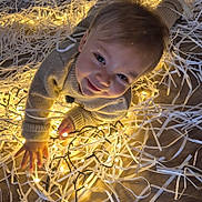 Gaspard participe au concours pour gagner de l'argent avec cette photo : toddler, child, smiling, fairy_lights, sweater, paper_shreds, curious, indoor, floor, decorative_lights, happy, young_child, cute, playful, cozy, warm_lighting, face, hand, wooden_floor, portrait