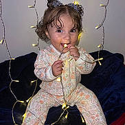 Athena is registered to the contest to win money with this photo: baby, child, infant, cute, smiling, lights, fairy_lights, glowing, hair_accessories, onesie, floral_pattern, sitting, indoor, blanket, playful, happy, portrait, cozy, festive, curly_hair