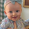 baby, child, smiling, bow, headband, hand, floral_clothing, portrait, indoor, person, face, happy, cute, infant, young_child, fingertips, fabric, decor, wall, holding