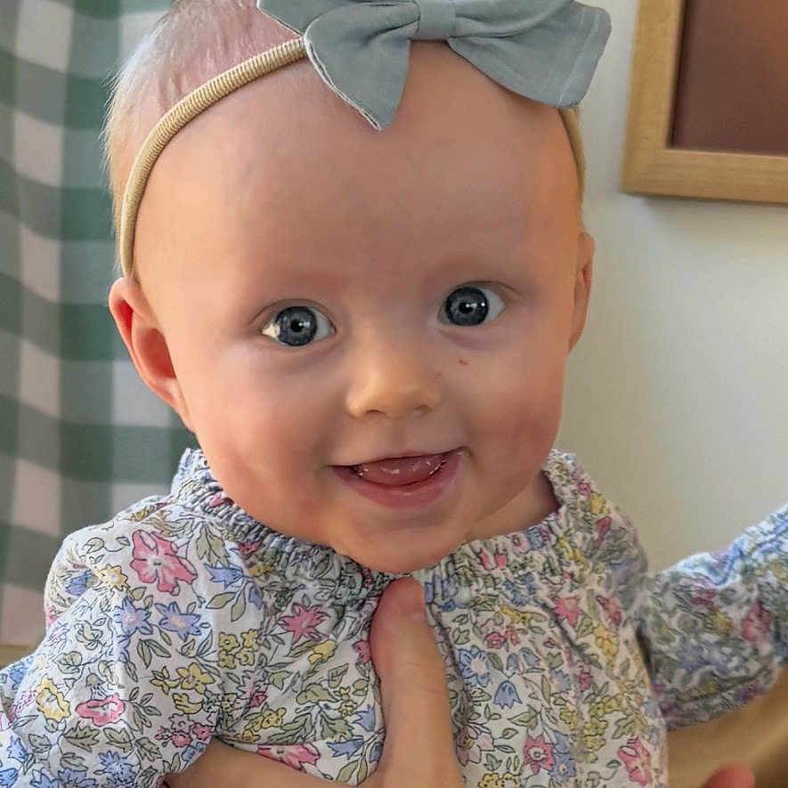 Sloane is registered to the contest to win money with this photo: baby, child, smiling, bow, headband, hand, floral_clothing, portrait, indoor, person, face, happy, cute, infant, young_child, fingertips, fabric, decor, wall, holding