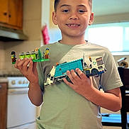 Cruz is registered to the contest to win money with this photo: child, boy, lego, toy, recycling_truck, mini_figures, kitchen, cabinet, window, light, smile, short_hair, green_shirt, camouflage_pants, indoor, person, holding, play, hobby, home
