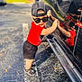 toddler, child, sunglasses, red_shirt, black_shorts, cap, reflection, car, outdoor, parking_lot, playful, sunny, footwear, sidewalk, shadow, person, cute, smiling, fashion, summer