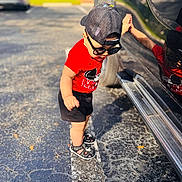 Bryan joined the competition — help win amazing prizes! toddler, child, sunglasses, cap, red_shirt, black_shorts, sandals, car, reflection, parking_lot, asphalt, line_marking, leaf, sunlight, shadow, outdoor, person, casual_clothing, summer, playful
