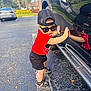 Bryan is registered to the contest to win money with this photo: toddler, child, sunglasses, cap, red_shirt, black_shorts, sandals, car, reflection, parking_lot, pavement, outdoor, daylight, trees, plants, shadow, cute, fashion, summer, side_view