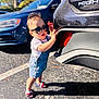 toddler, child, sunglasses, denim_overalls, car, parking_lot, shadow, sunlight, asphalt, summer, footwear, sandals, exploring, outdoor, person, vehicle, casual, cute, fashion, daytime