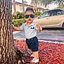 toddler, child, tree, sunglasses, shorts, shirt, shoes, outdoor, daylight, car, sidewalk, mulch, greenery, residential, person, standing, fashion, cute, hairstyle, summer
