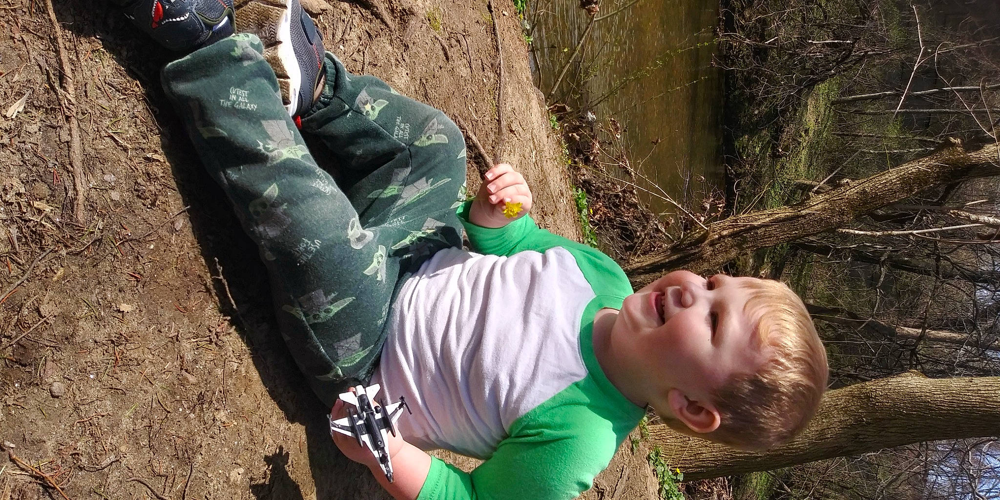 Luke is registered to the contest to win money with this photo: adventure, camouflage, child, forest, fun, grass, human_body, leisure, military_camouflage, people_in_nature, person, recreation, rock, sitting, sneakers, soil, t_shirt, tree, trunk, wood