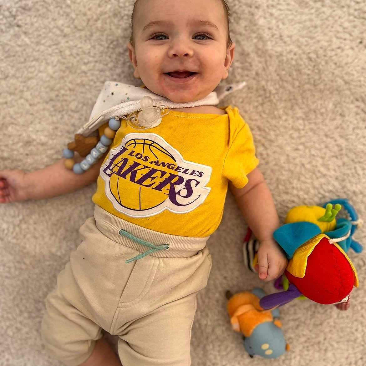Lyam participe au concours pour gagner de l'argent avec cette photo : baby, infant, smiling, yellow_shirt, los_angeles_lakers, pacifier, plush_toy, carpet, beige_pants, child, happy, lying_down, cute, indoors, casual_clothing, colorful_toys, young_child, face, small_hands, playful