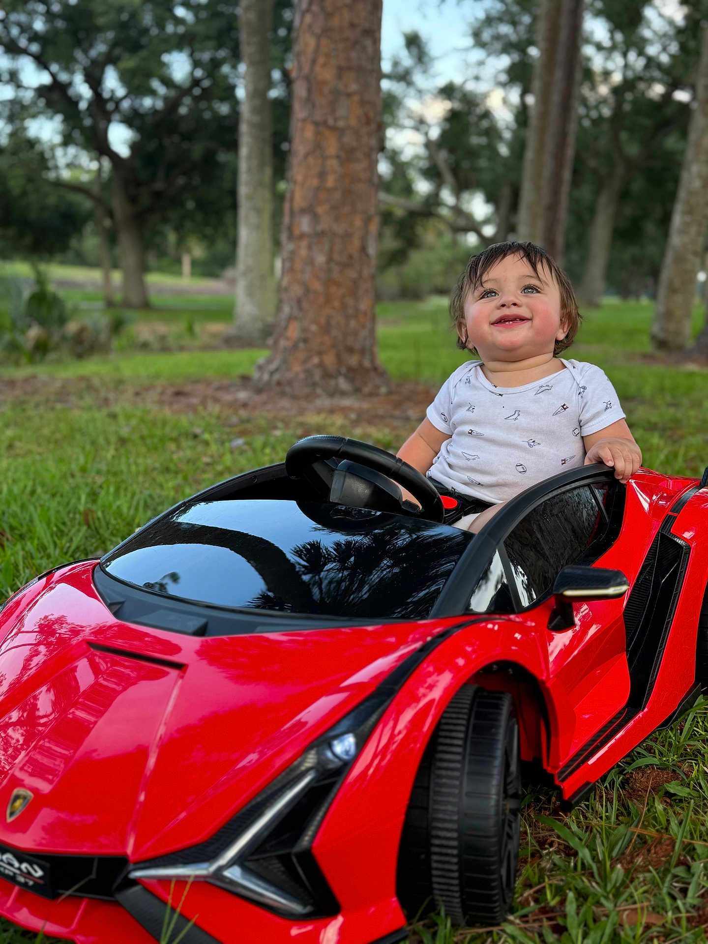 Yazan joined the competition — help win amazing prizes! child, toddler, toy_car, red_car, outdoor, grass, trees, greenery, happy, smiling, baby, park, nature, play, cute, vehicle, steering_wheel, daylight, casual_clothing, fun
