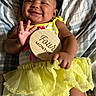 baby, smile, yellow_dress, wooden_sign, four_months, hand, face, blanket, pattern, infant, happy, lying_down, cute, clothing, child, portrait, celebration, milestone, small_hand, indoors