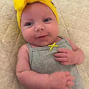 Oaklee joined the competition — help win amazing prizes! baby, infant, headband, yellow, bow, grey_outfit, cute, smiling, lying_down, pale_skin, hand, curious, closeup, indoors, quilted_surface, child, small_ribbon, soft_light, portrait, adorable