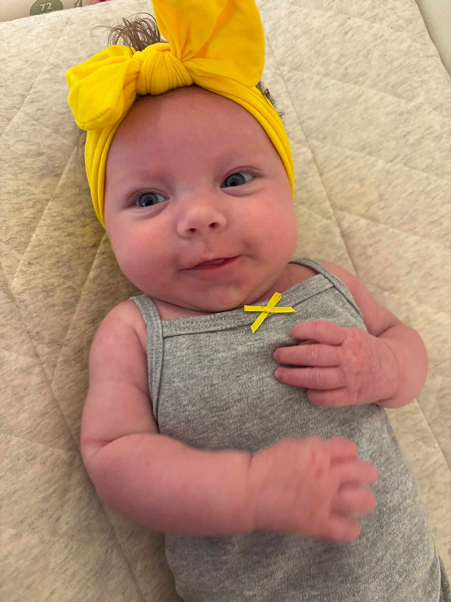 Oaklee joined the competition — help win amazing prizes! baby, infant, headband, yellow, bow, grey_outfit, cute, smiling, lying_down, pale_skin, hand, curious, closeup, indoors, quilted_surface, child, small_ribbon, soft_light, portrait, adorable