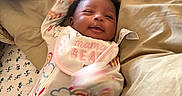 Starr is registered to the contest to win money with this photo: baby, infant, child, smiling, portrait, lying_down, bed, bedding, pillows, blanket, onesie, bib, rainbows, clouds, colorful_clothing, indoors, bedroom, cozy, happy, face