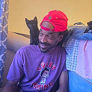 Ocean is registered to the contest to win money with this photo: man, black_kitten, cat, red_cap, purple_tshirt, smiling, indoor, curtain, bed, clothes, shoulder, facial_hair, casual, seated, jeans, patterned, looking_at_camera, cozy, young_adult, friendly