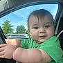 baby, child, car, steering_wheel, green_shirt, curious_expression, chubby_cheeks, window, sky, trees, daylight, portrait, person, seat, hand, arm, indoors, casual_clothing, vehicle, driver
