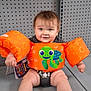 baby, child, floaties, orange, turtle, smiling, sitting, pegboard, gray, metal, shelf, cute, happy, infant, clothing, toy, indoor, person, legs, feet