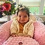 baby, smile, pink_cushion, bow, hair, flower_vase, flowers, window, table, indoor, child, cute, happy, infant, clothing, jacket, bracelet, portrait, cozy, home
