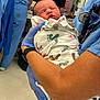 newborn, baby, hospital, medical_staff, blue_gloves, blanket, footprints, scrubs, healthcare_worker, patient_care, infant, holding, indoor, medical_equipment, person, clinic, face, sleeping, close_up, care