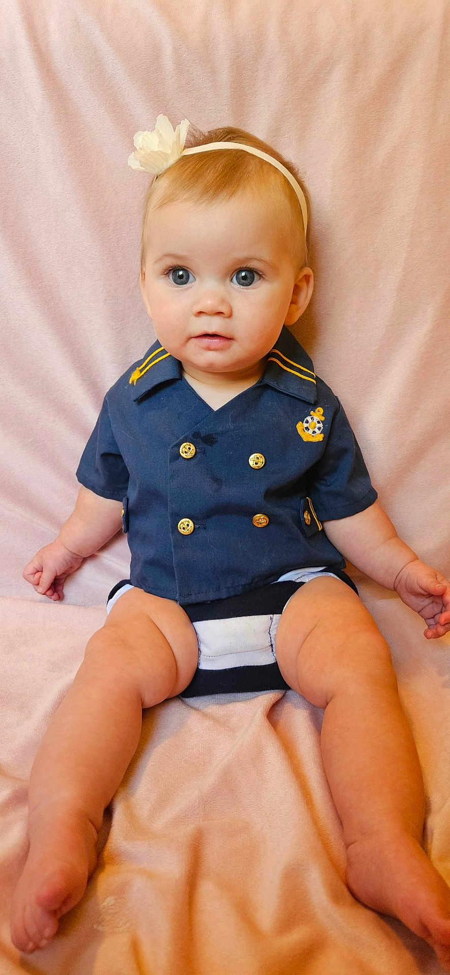 Kaston is registered to the contest to win money with this photo: baby, child, infant, sailor_shirt, blue_eyes, flower_headband, sitting, pink_blanket, cute, portrait, clothing, gold_buttons, soft_texture, indoors, skin, person, legs, hands, head, face
