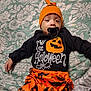 William is registered to the contest to win money with this photo: baby, child, halloween, costume, pumpkin, orange, black, pacifier, hat, clothing, lying_down, bedspread, floral_pattern, infant, cute, holiday, celebration, indoors, person, smiling