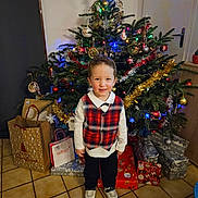 Noa participe au concours pour gagner de l'argent avec cette photo : child, boy, christmas_tree, presents, gift_bags, holiday, festive_clothing, plaid_vest, white_shirt, sneakers, tiled_floor, decorations, tinsel, christmas_lights, indoor, smiling, holiday_season, celebration, festive, home