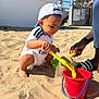 adult_hand, beach, blue_sky, bucket, building, cap, child, footwear, fun, outdoor, people, playing, recreation, sand, shovel, summer, sunny, toddler, vacation, white_clothing