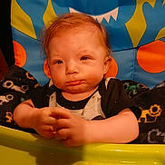 Sebastian joined the competition — help win amazing prizes! baby, child, high_chair, colorful, monster_design, food, hands, face, expression, indoor, clothing, blanket, seat, person, cute, infant, orange, blue, green, yellow
