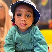 El Mehdi is registered to the contest to win money with this photo: toddler, child, face, hat, bucket_hat, sweatshirt, blue, teal, curious, indoors, portrait, person, big_eyes, expression, young_child, casual_clothing, blurred_background, yellow_chair, soft_lighting, close_up