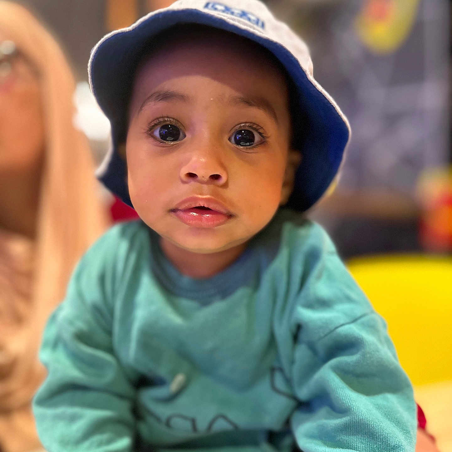 El Mehdi is registered to the contest to win money with this photo: big_eyes, blue, blurred_background, bucket_hat, casual_clothing, child, close_up, curious, expression, face, hat, indoors, person, portrait, soft_lighting, sweatshirt, teal, toddler, yellow_chair, young_child