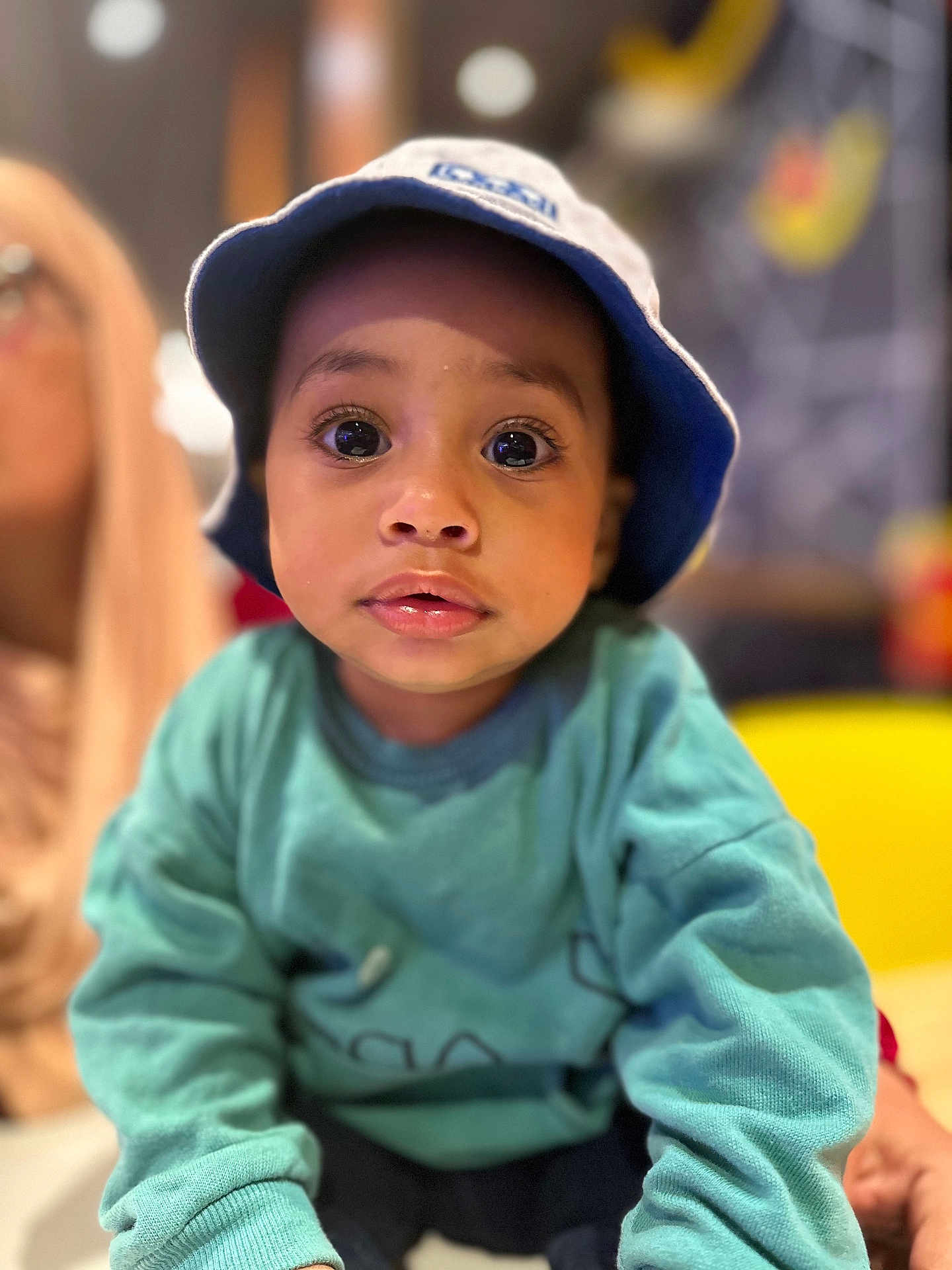 El Mehdi is registered to the contest to win money with this photo: toddler, child, face, hat, bucket_hat, sweatshirt, blue, teal, curious, indoors, portrait, person, big_eyes, expression, young_child, casual_clothing, blurred_background, yellow_chair, soft_lighting, close_up