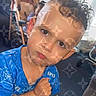 child, boy, curly_hair, blue_shirt, serious_expression, indoor, furniture, background_child, pattern_overlay, portrait, close_up, face, young_child, casual_clothing, home, person, expression, sitting, room, hair