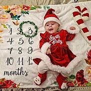 Elena a rejoint le concours — aidez-le/la à gagner de superbes lots ! baby, infant, christmas, holiday, santa_hat, santa_baby, red_dress, milestone_blanket, months, floral_pattern, candy_cane, plush_toy, socks, smiling, lying_down, green_wreath, cute, festive, celebration, indoors