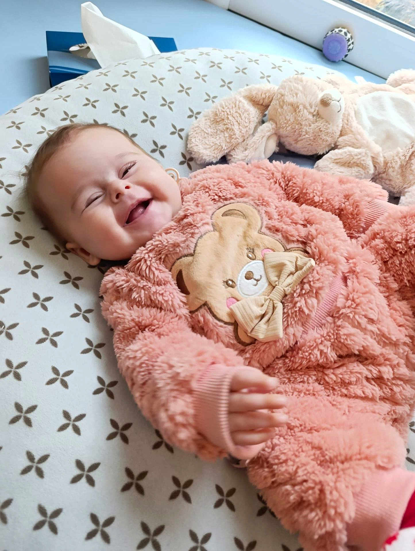 Elena participe au concours pour gagner de l'argent avec cette photo : baby, child, smiling, pink_clothing, plush_toy, stuffed_animal, cushion, patterned_bedding, indoor, happy, infant, onesie, soft_texture, cute, reaching_hand, face, head, window, light, cozy