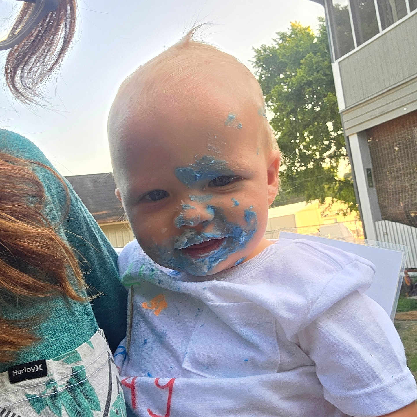 Robert Michels joined the competition — help win amazing prizes! baby, child, infant, face, smiling, frosting, blue_cream, messy_face, white_shirt, outdoor, sunlight, portrait, being_held, adult, house, trees, backyard, summer, closeup, funny
