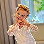 blurry_background, child, curtains, cute, flower_crown, girl, happy, headwear, heart_hands, indoor, lace_clothing, person, portrait, posing, smile, soft_lighting, toddler, warm_colors, window, young_child