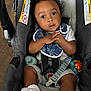 baby, child, car_seat, safety_harness, bib, curly_hair, white_socks, toy, floor, wood_flooring, infant, seat_belt, expression, hands, clothing, indoor, warning_label, face, portrait, person