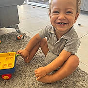Loan a rejoint le concours — aidez-le/la à gagner de superbes lots ! toddler, child, smiling, curly_hair, grey_polo, onesie, toy_cart, sitting, rug, carpet, living_room, indoor, happy, play, furniture, floor, tile, cute, person, home