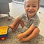 toddler, child, smiling, curly_hair, grey_polo, onesie, toy_cart, sitting, rug, carpet, living_room, indoor, happy, play, furniture, floor, tile, cute, person, home