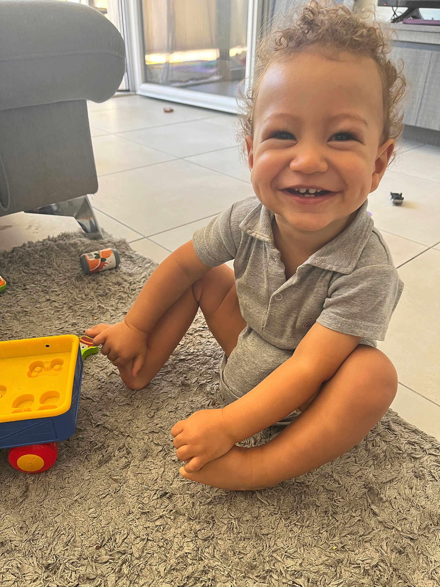 Loan a rejoint le concours — aidez-le/la à gagner de superbes lots ! toddler, child, smiling, curly_hair, grey_polo, onesie, toy_cart, sitting, rug, carpet, living_room, indoor, happy, play, furniture, floor, tile, cute, person, home