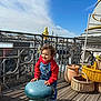 toddler, child, outdoor, terrace, sky, clouds, railing, stool, overalls, hoodie, smile, curly_hair, wicker_basket, clay_pot, wooden_floor, building, urban, daylight, eiffel_tower, happy