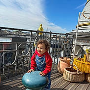 Sam participe au concours pour gagner de l'argent avec cette photo : toddler, child, outdoor, terrace, sky, clouds, railing, stool, overalls, hoodie, smile, curly_hair, wicker_basket, clay_pot, wooden_floor, building, urban, daylight, eiffel_tower, happy