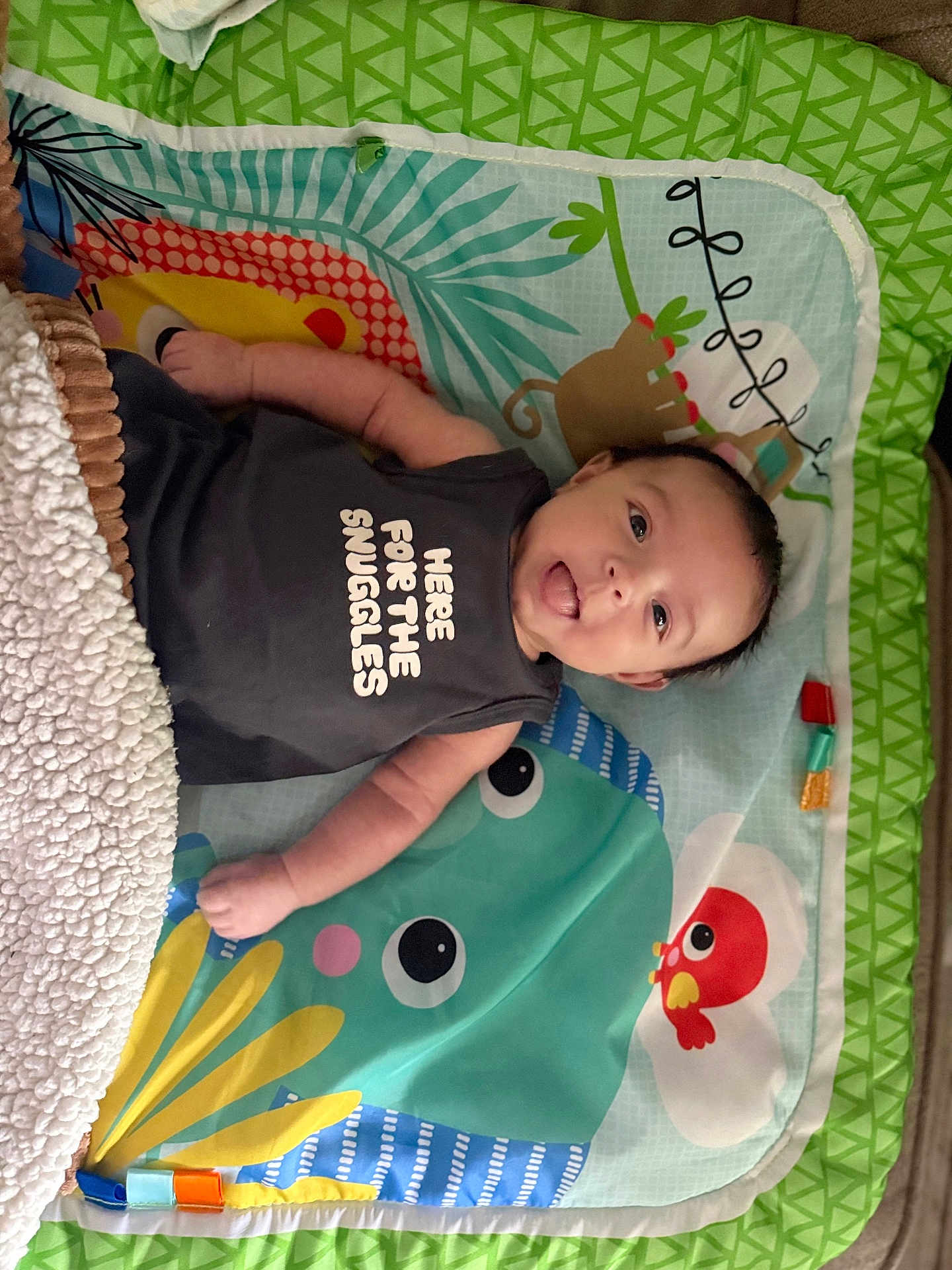 Lucas is registered to the contest to win money with this photo: baby, infant, face, smile, tongue, onesie, text_on_clothing, blanket, playmat, toy, cartoon_animal, colorful_pattern, plush, arm, hand, eyes, hair, indoor, happy, portrait