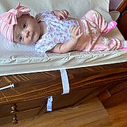 Rayna is registered to the contest to win money with this photo: baby, infant, pink_clothing, headband, changing_pad, wooden_dresser, floor, hand, face, eyes, cute, indoor, child, newborn, apparel, fabric, soft_lighting, room, person, portrait