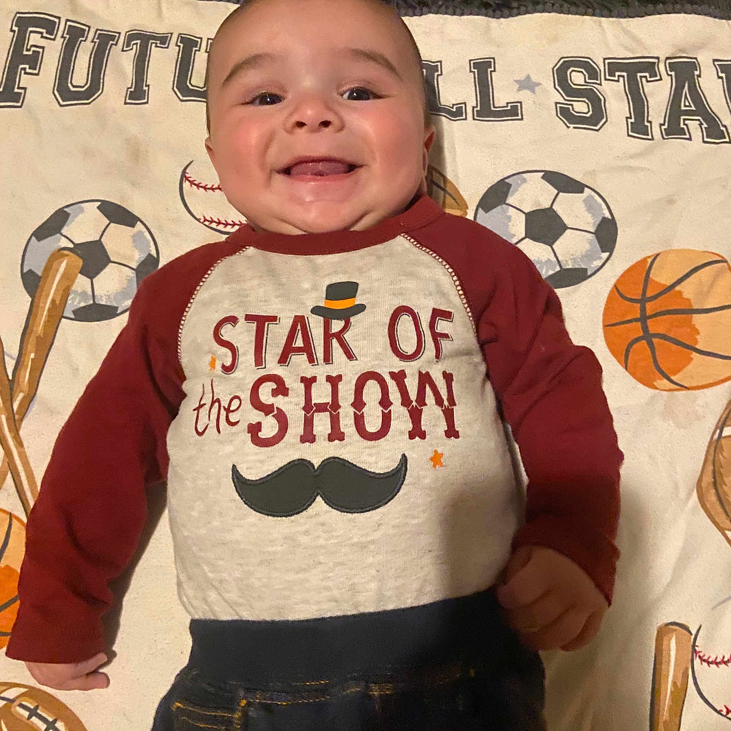 Ezra joined the competition — help win amazing prizes! baby, smiling, infant, clothing, blanket, sports, baseball, soccer_ball, basketball, mustache_graphic, shirt, cute, happy, lying_down, child, portrait, indoors, young_child, playful, face