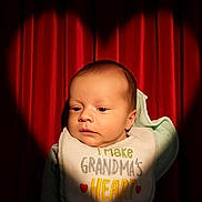 Sillis is registered to the contest to win money with this photo: baby, infant, newborn, bib, red_curtain, heart_spotlight, portrait, face, head, eyes_open, warm_lighting, cute, adorable, clothing, indoor, close_up, shadow, fabric, sitting, calm