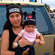Nala is registered to the contest to win money with this photo: woman, baby, hat, black_hat, text_on_hat, outdoor, car, vehicle, person, child, holding, cute, dusk, casual_clothing, toy, pink_bib, smile, face, portrait, family