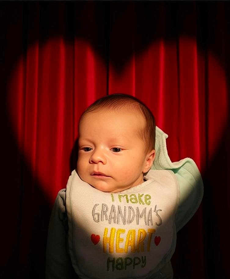 Sillis joined the competition — help win amazing prizes! baby, infant, newborn, bib, red_curtain, heart_spotlight, portrait, face, head, eyes_open, warm_lighting, cute, adorable, clothing, indoor, close_up, shadow, fabric, sitting, calm