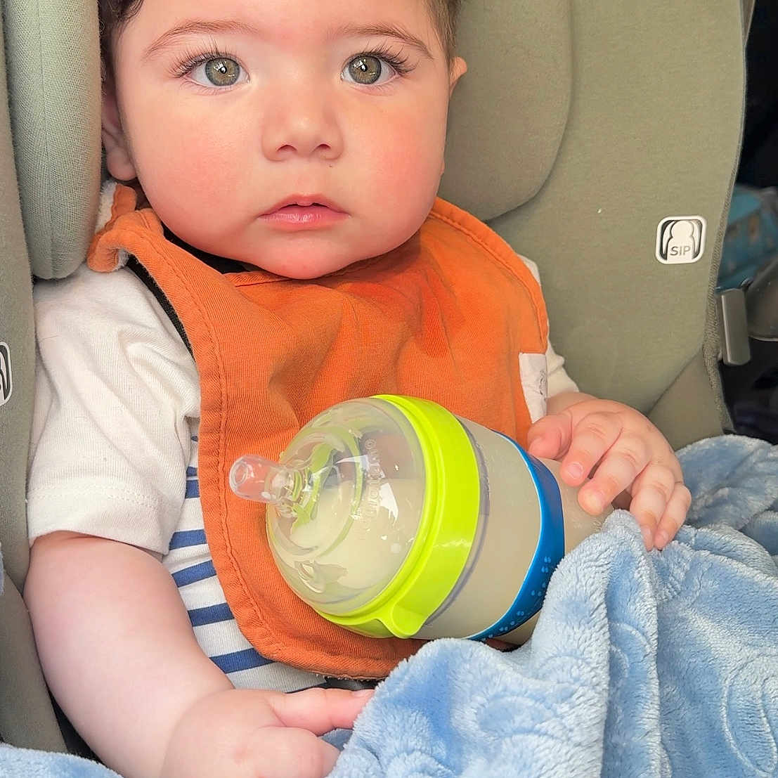 Sameh is registered to the contest to win money with this photo: baby, blanket, blue_blanket, bottle, car, car_seat, child, close_up, curly_hair, cute, feeding_bottle, holding, indoor, infant, orange_bib, person, portrait, seat, soft_texture, young_child